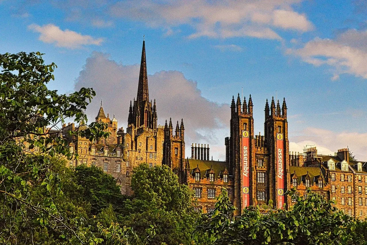 Historische Steingebäude und ein hoher, spitzer Turm ragen über grünen Bäumen in Edinburgh empor. Rote Banner mit 'assembl...