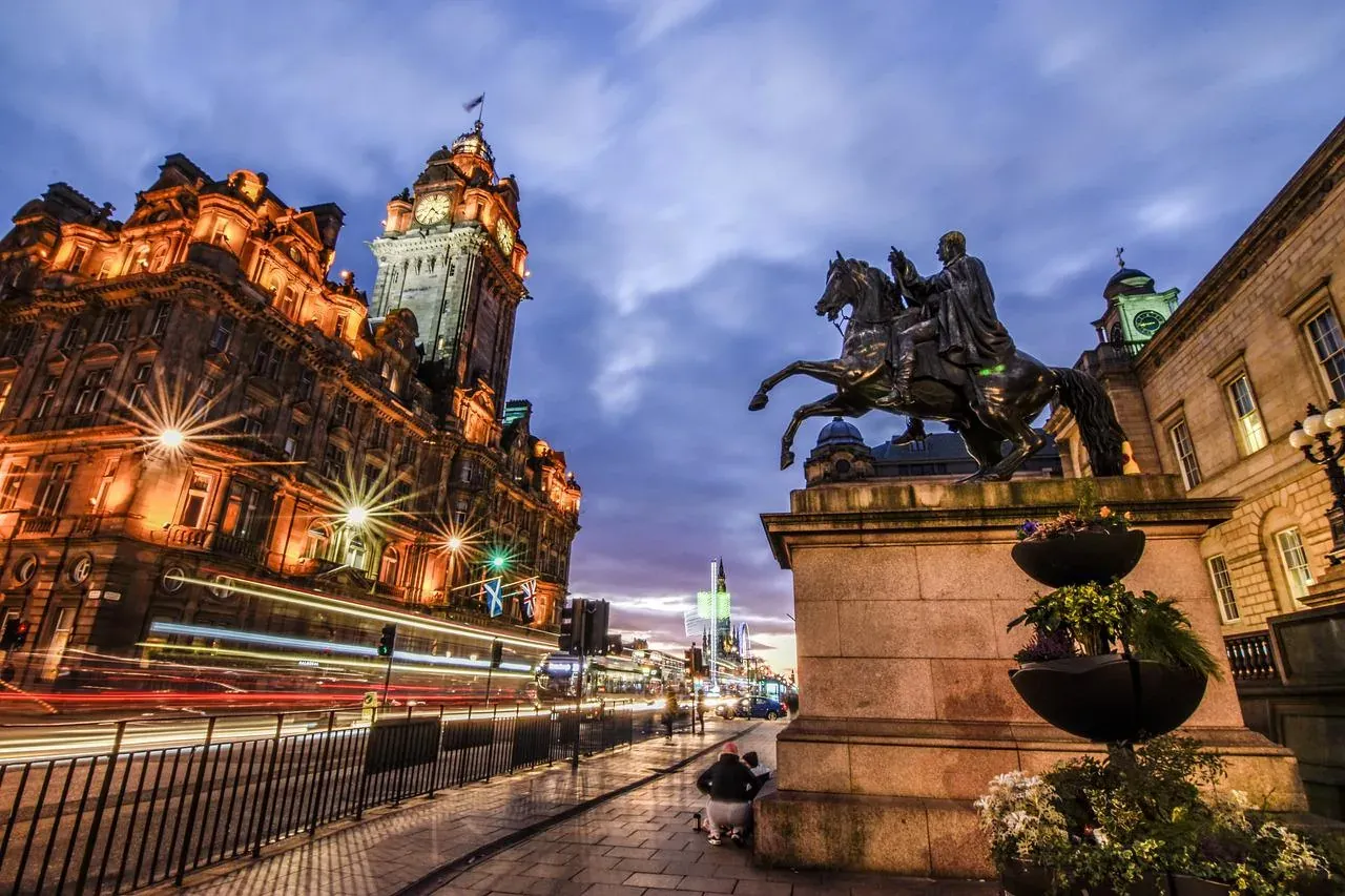 Beleuchteter Uhrturm und Fassade des Balmoral Hotels bei Dämmerung, mit Reiterstatue, Straßenlaternen und Lichtspuren von ...
