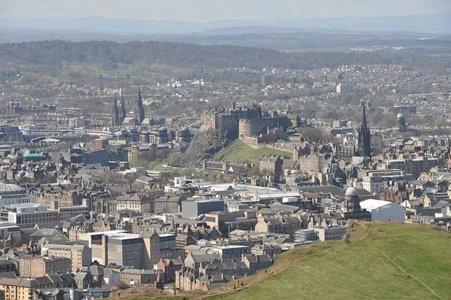 Panoramablick auf Edinburgh von einem Hügel, mit Edinburgh Castle auf einem Felsen, Stadtgebäuden und fernen grünen Hügeln.