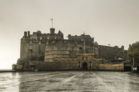 Die imposante Steinfassade und Zinnen von Edinburgh Castle unter bedecktem Himmel. Die nasse Esplanade im Vordergrund refl...