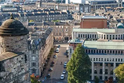 Blick auf Edinburghs Stadtbild von einem erhöhten Punkt, mit einem steinernen Burgturm links, einer Straße und fernen Hügeln.