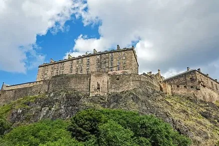 Edinburgh Castle auf einem felsigen Hügel mit grünen Bäumen davor, unter einem teilweise bewölkten blauen Himmel.