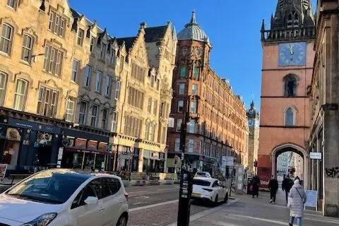 Historic street in Glasgow with ornate stone buildings, a prominent reddish-orange clock tower, and a white car parked on ...