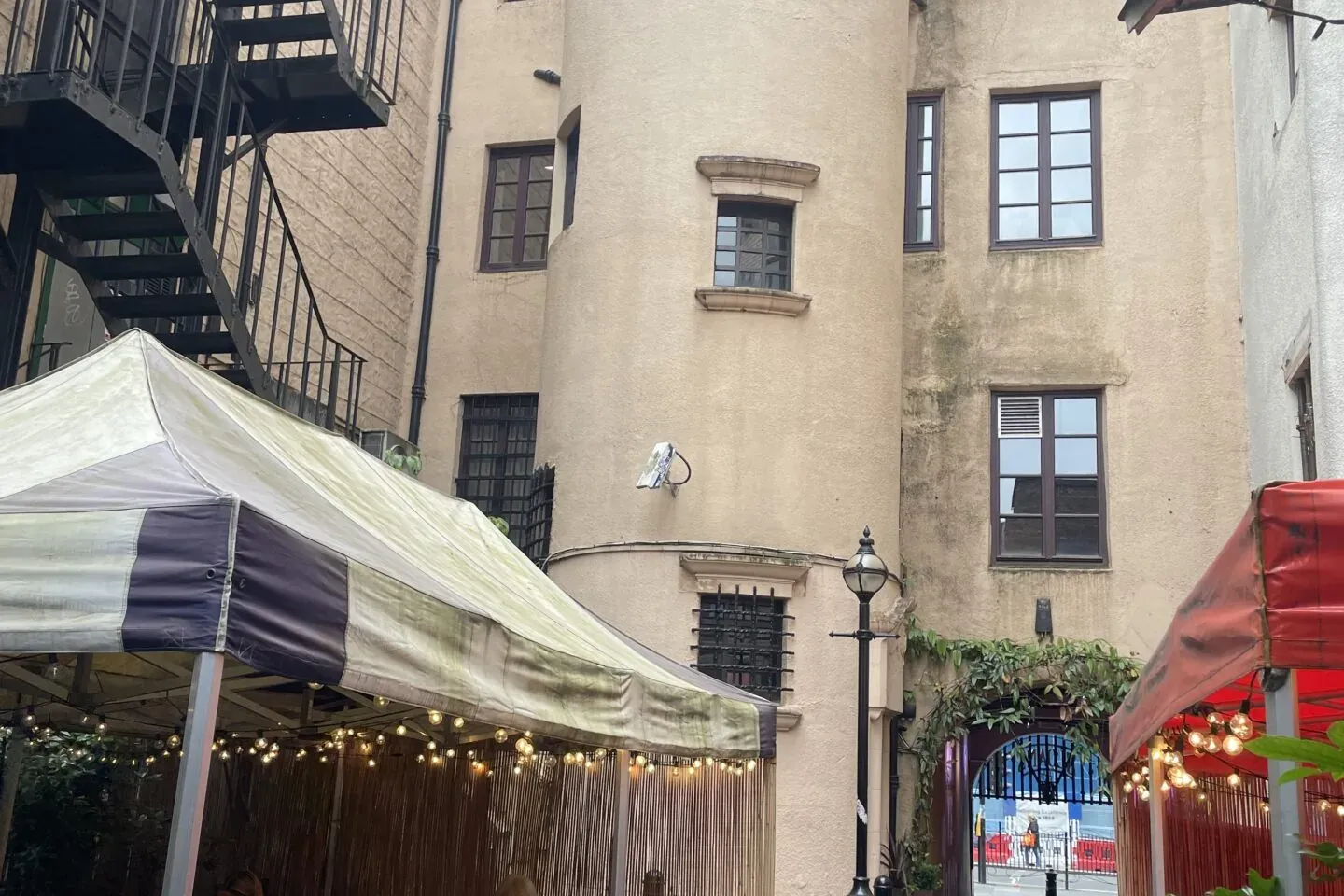 Personas cenando en una cafetería al aire libre bajo toldos de rayas en un estrecho callejón empedrado, rodeado de edifici...