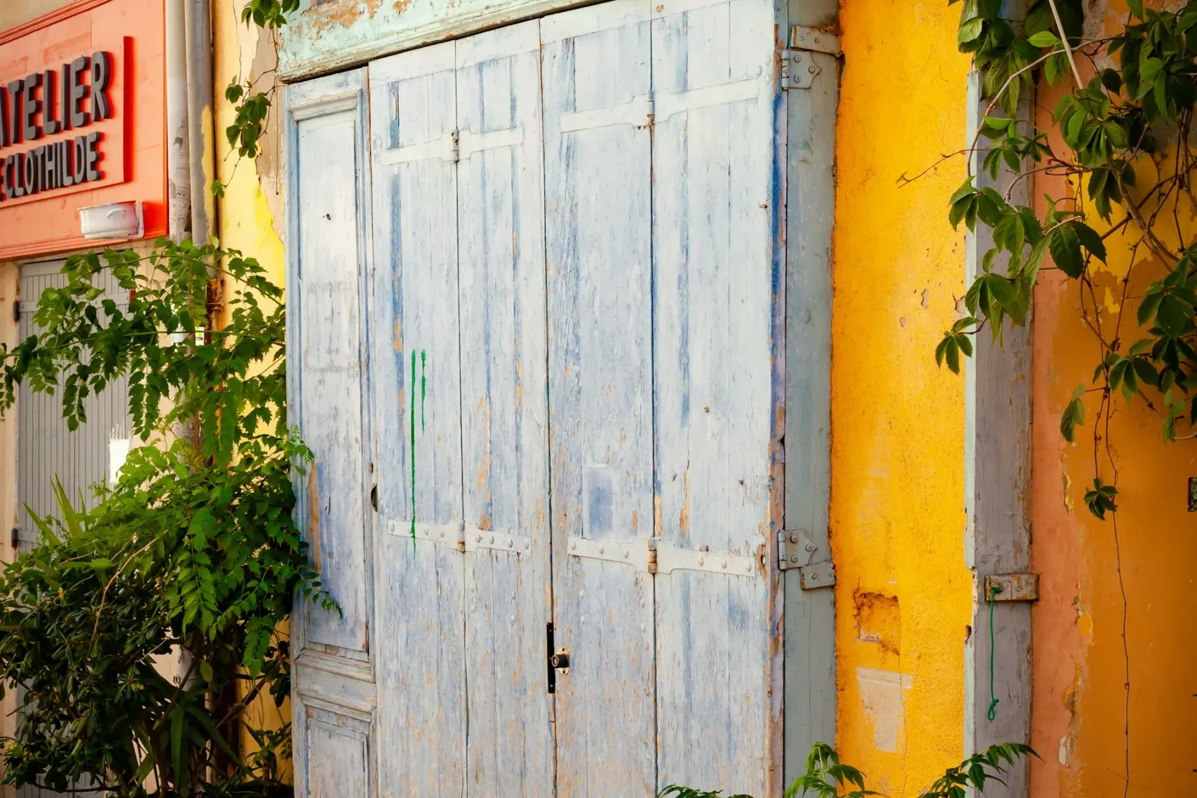 Puertas de madera azul claro desgastadas de una tienda con letrero 'Bazar du Panier', paredes amarillas y enredaderas verdes.