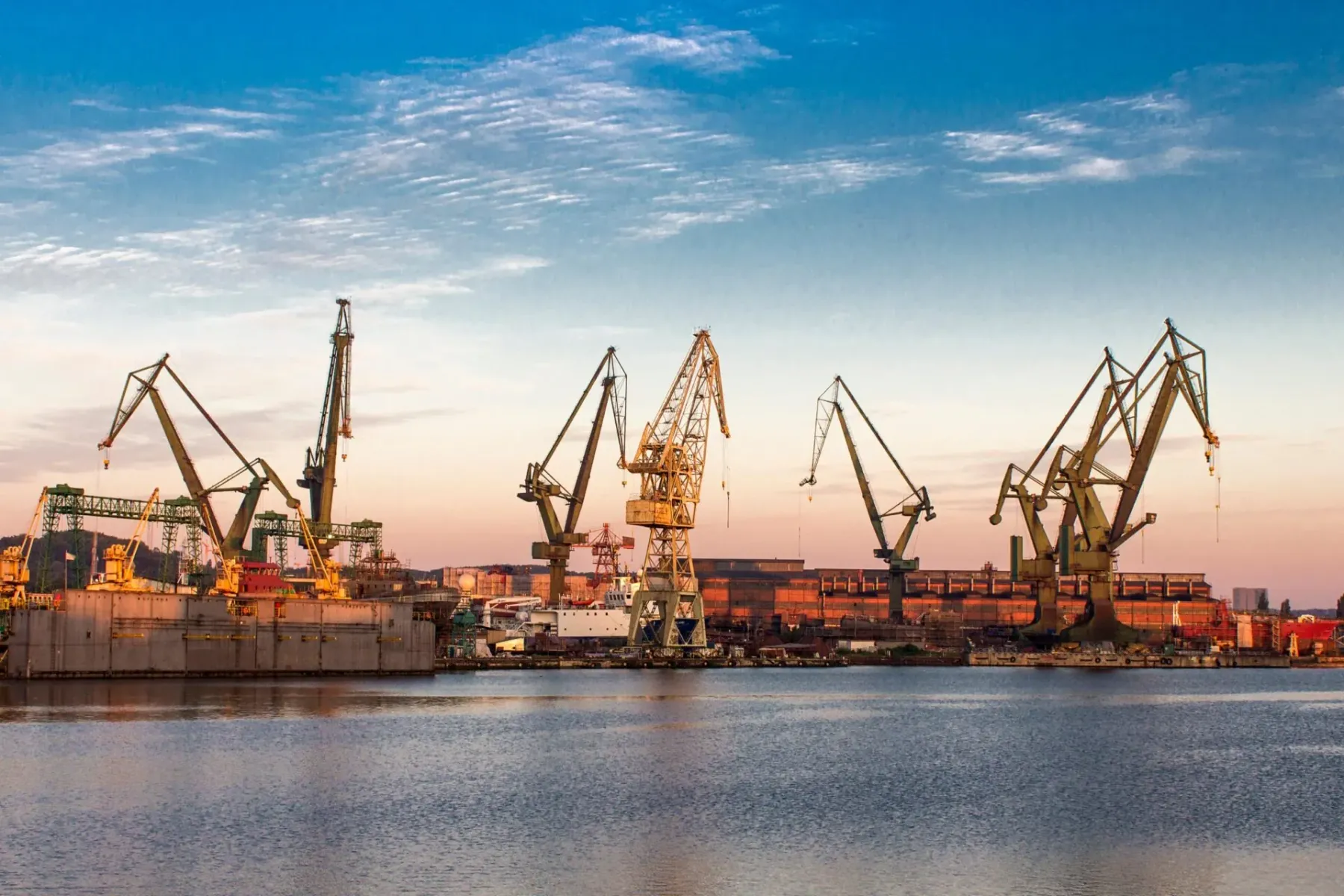 Mehrere große Industriekräne und Werftgebäude unter einem teilweise bewölkten Himmel, mit Spiegelungen im Wasser bei Sonne...