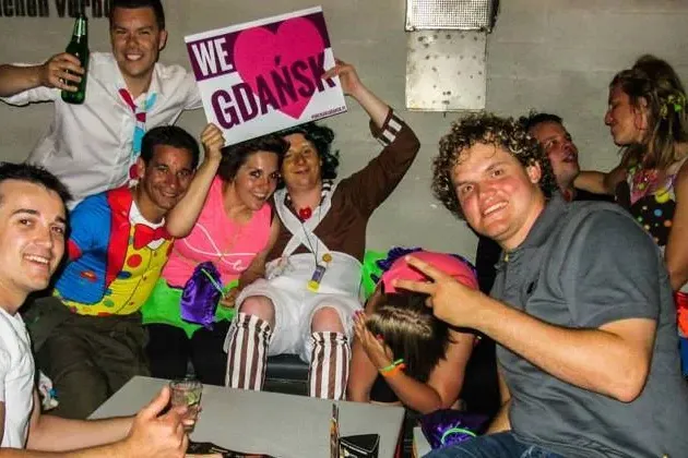 Gruppe lächelnder Personen, teils in Kostümen, die ein 'We <3 Gdańsk' Schild in Innenräumen mit Getränken halten.