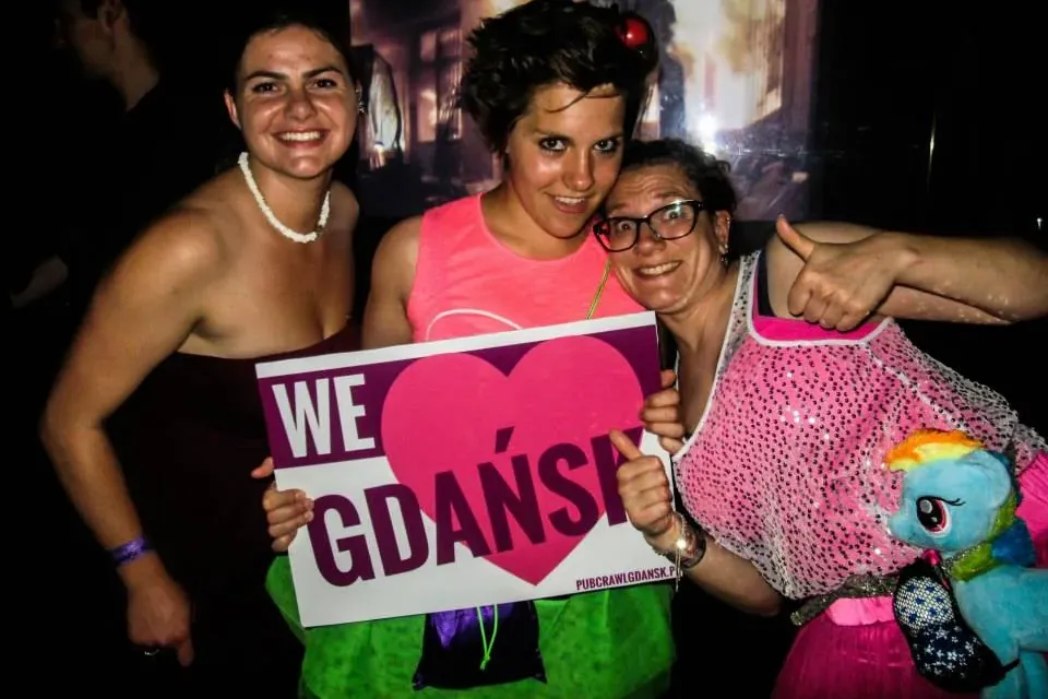 Drei lächelnde Frauen posieren mit einem weißen Schild 'WE ❤️ GDAŃSK' und 'pubcrawlgdansk.pl' vor dunklem Hintergrund.