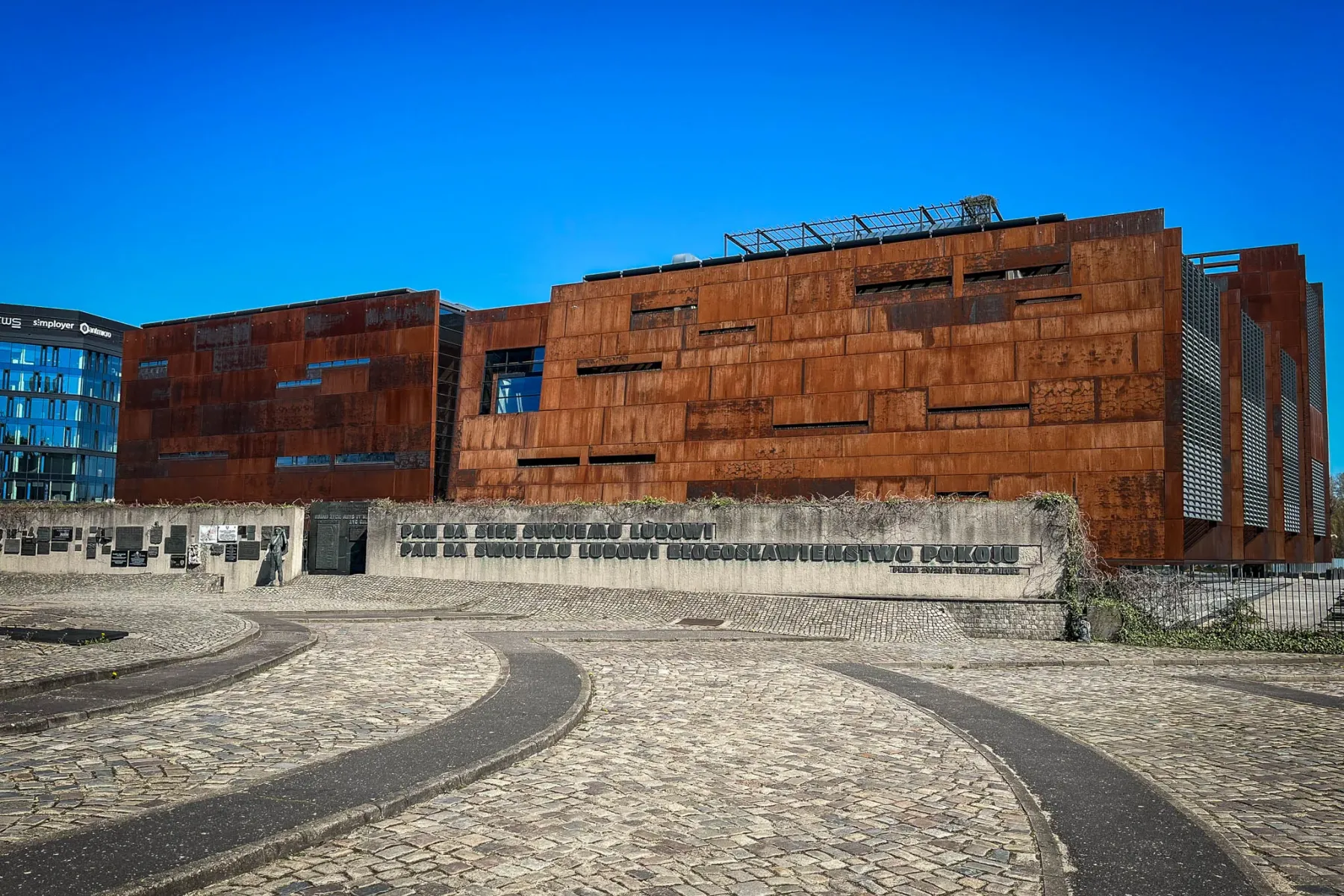 Gebäude mit verrosteter Metallfassade, gepflastertem Vorplatz und Beton-Gedenkmauer mit Tafeln und polnischer Schrift. Bla...