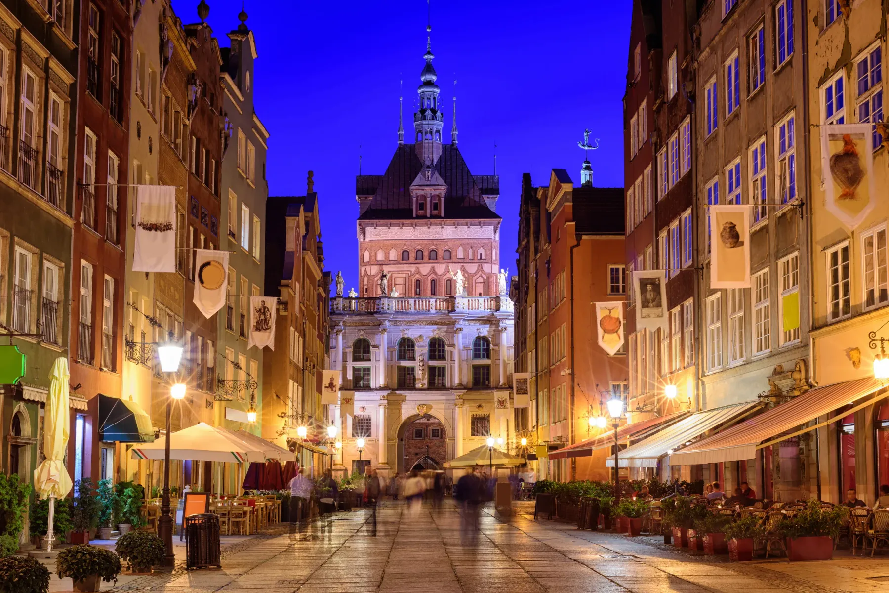 La calle Długa de Gdańsk al anochecer, con edificios históricos y cafeterías al aire libre. El Ayuntamiento iluminado al f...
