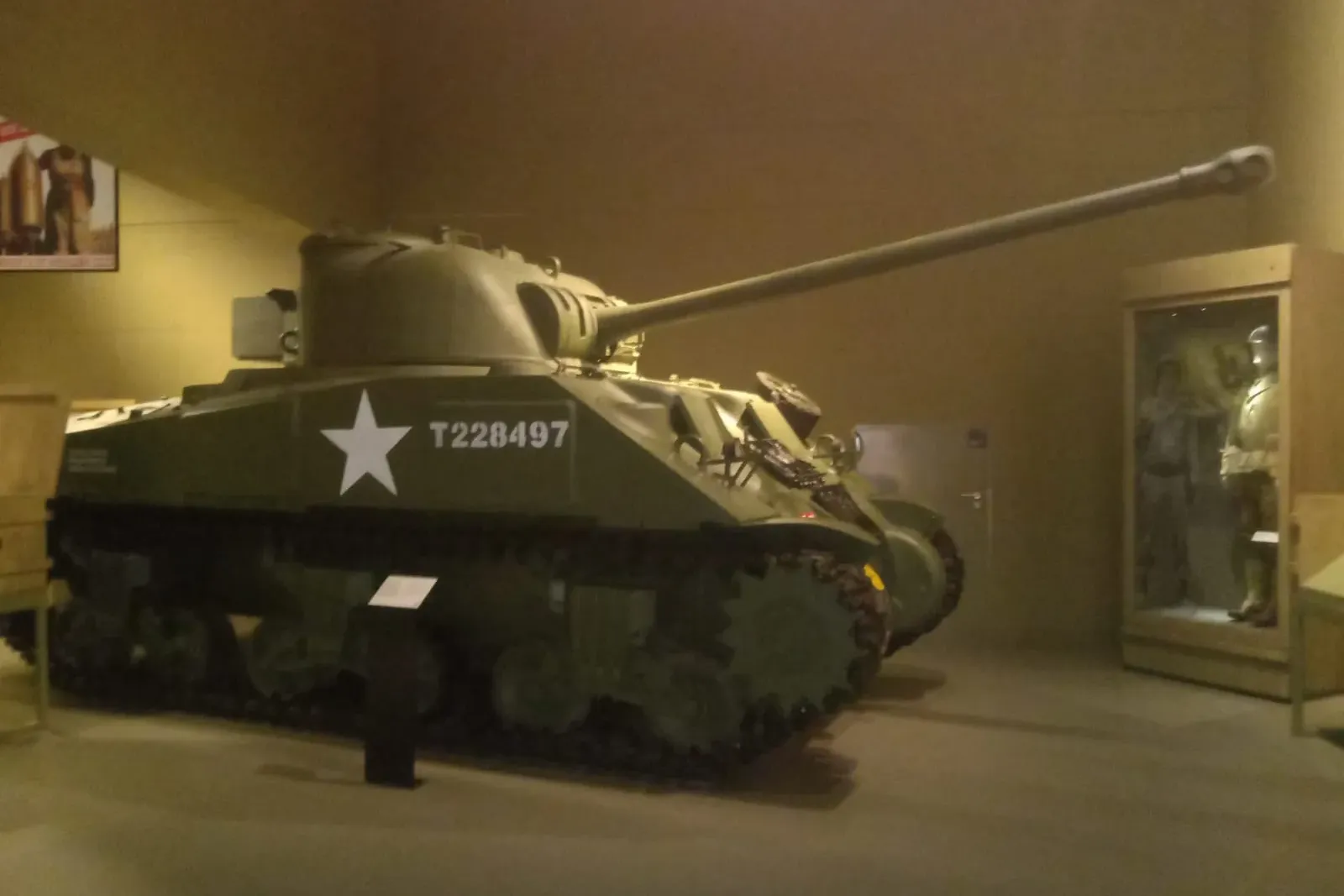 Grüner Sherman M4 Panzer mit weißem Stern und T228497 an der Seite, ausgestellt in einem schwach beleuchteten Museumssaal.