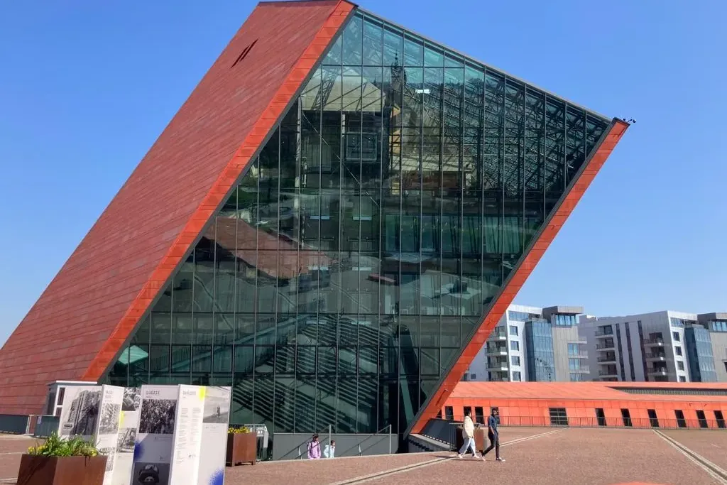 Modernes Gebäude mit einer großen, schrägen, rotbraunen Wand und reflektierender Glasfassade, Menschen auf einem gepflaste...