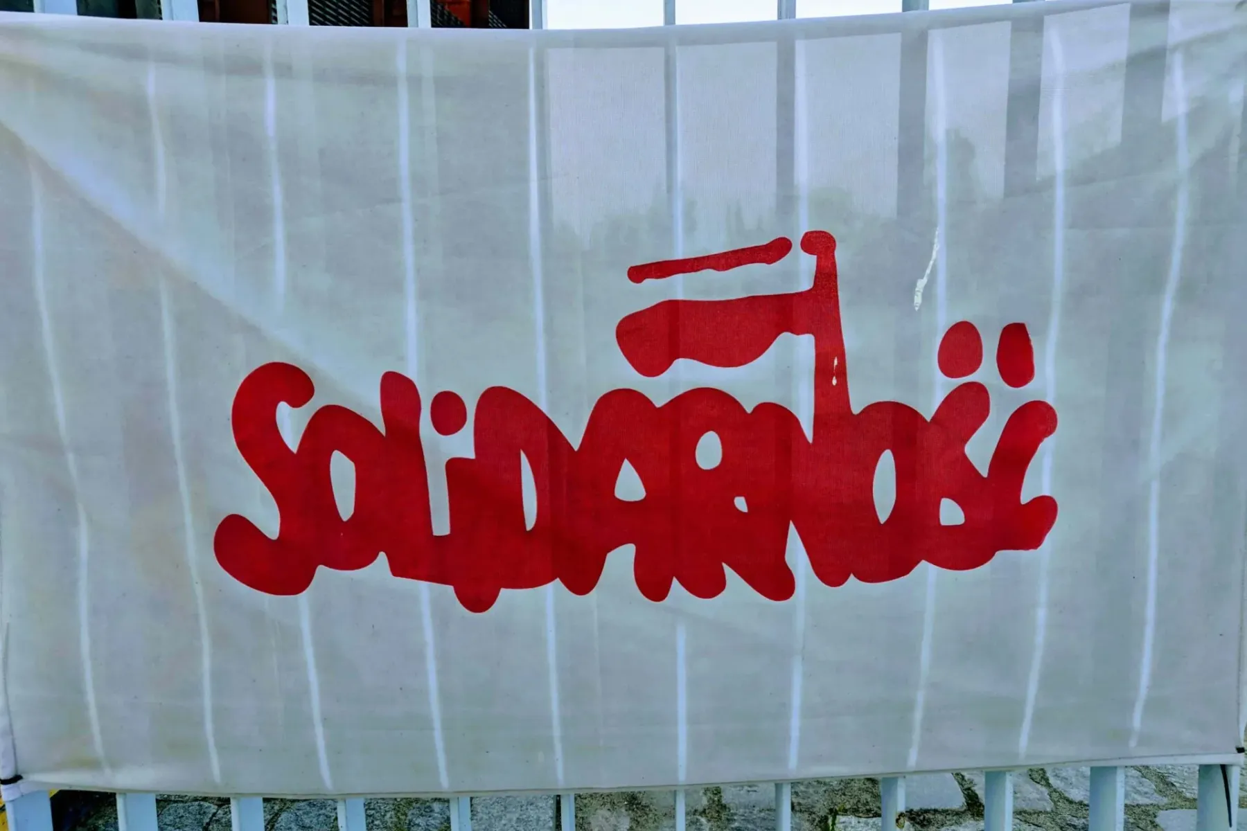 Weißes Banner mit rotem Solidarność-Logo, das eine stilisierte polnische Flagge zeigt. Das Banner hängt an einem weißen Me...
