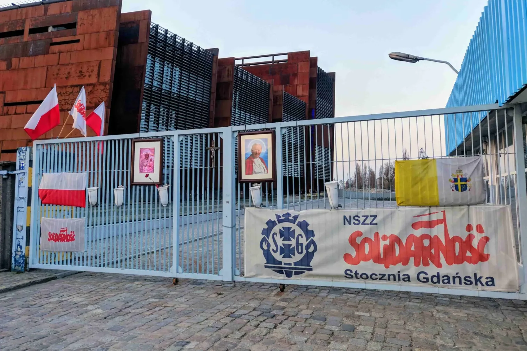 Metalltor mit polnischen und vatikanischen Flaggen, Solidarność-Bannern und gerahmten Bildern vor einem rostfarbenen Gebäude.