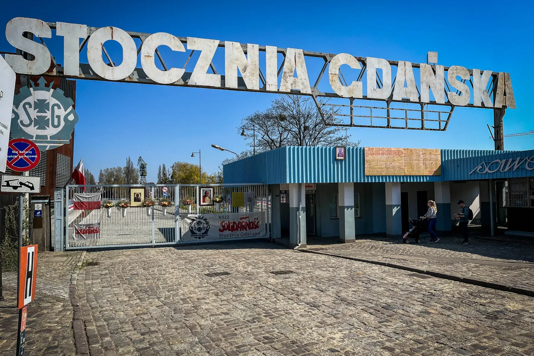 Großes Metallschild "STOCZNIA GDAŃSKA" über einem Tor mit Solidarność-Banner und polnischen Flaggen, Kopfsteinpflaster.