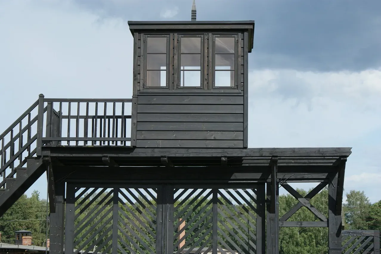 Dunkler Holzwachturm mit kleinem Raum und drei Fenstern, Treppe und Lattenzaun davor, vor bewölktem Himmel.