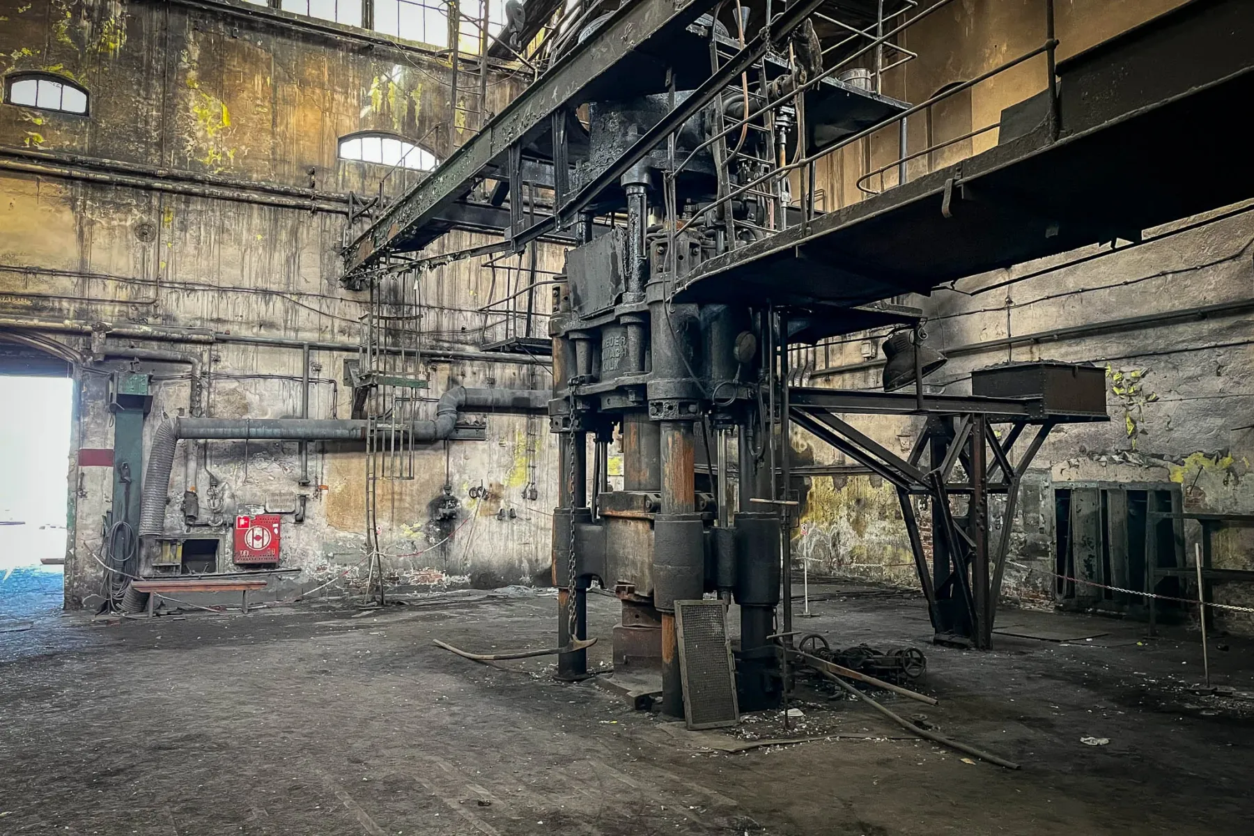 Verlassene Industriehalle mit großer, dunkler Hydraulikpresse im Zentrum, schmutzigem Boden, abblätternden Wänden und hohe...