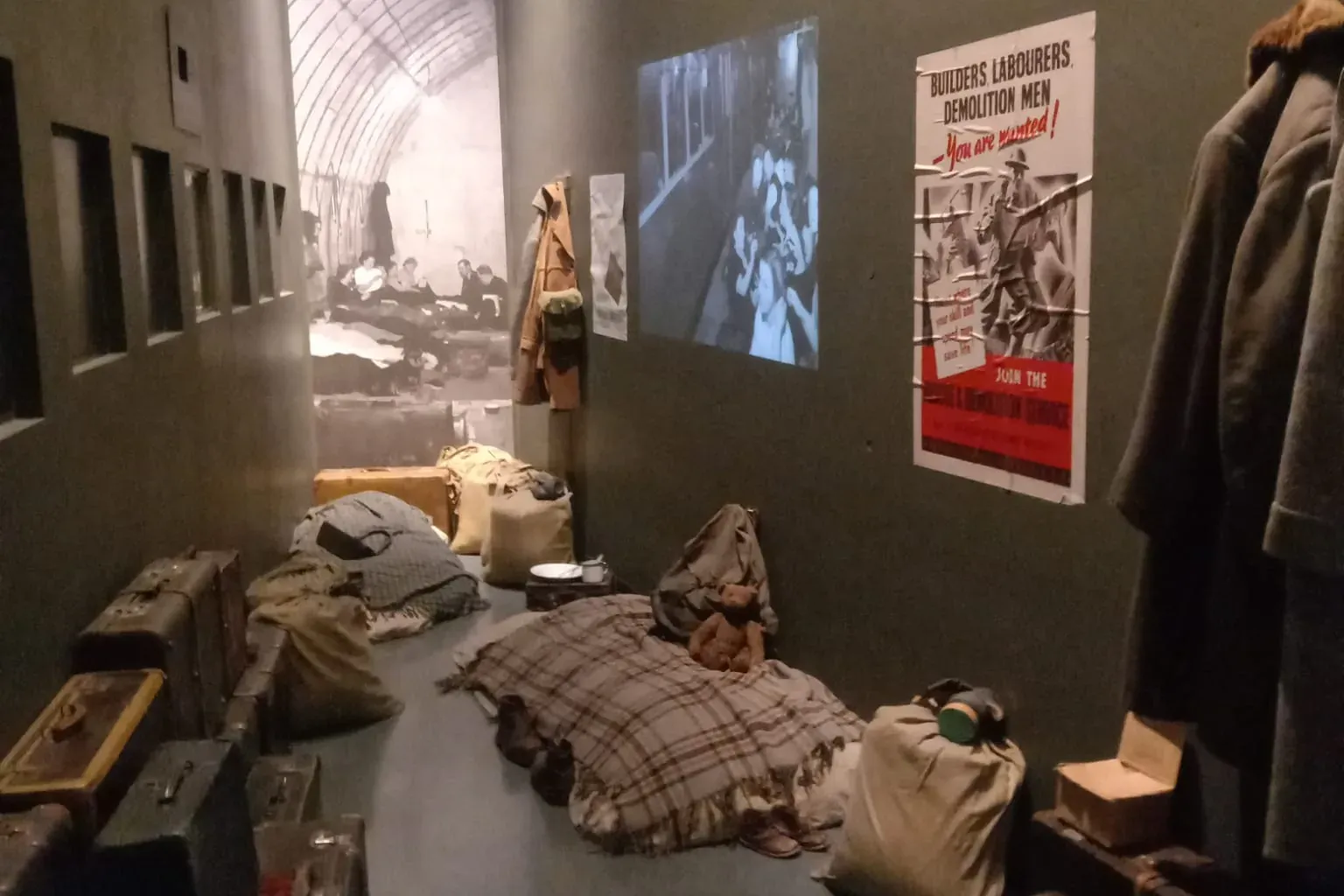 Dunkler Museumsraum, der einen Kriegsbunker zeigt, mit gestapelten Koffern, zerwühlten Decken, Kleidung und einem Propagan...
