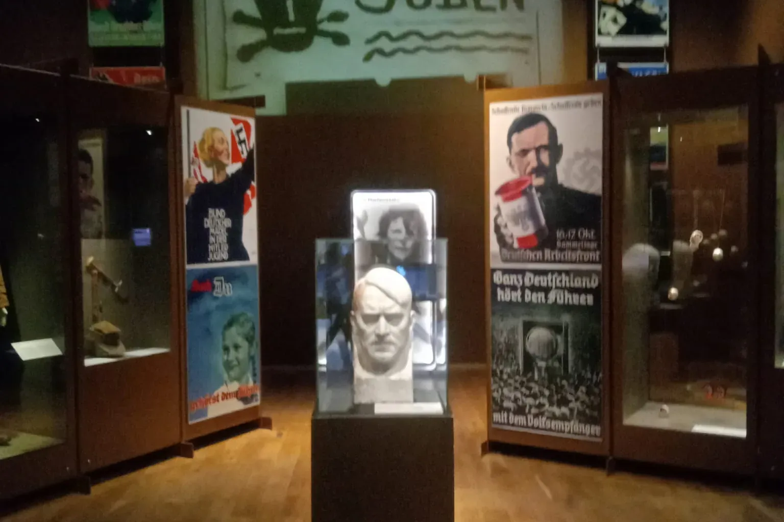 Museumsausstellung mit einer Büste auf einem Sockel, umgeben von Vitrinen und historischen Propagandaplakaten, darunter 'A...