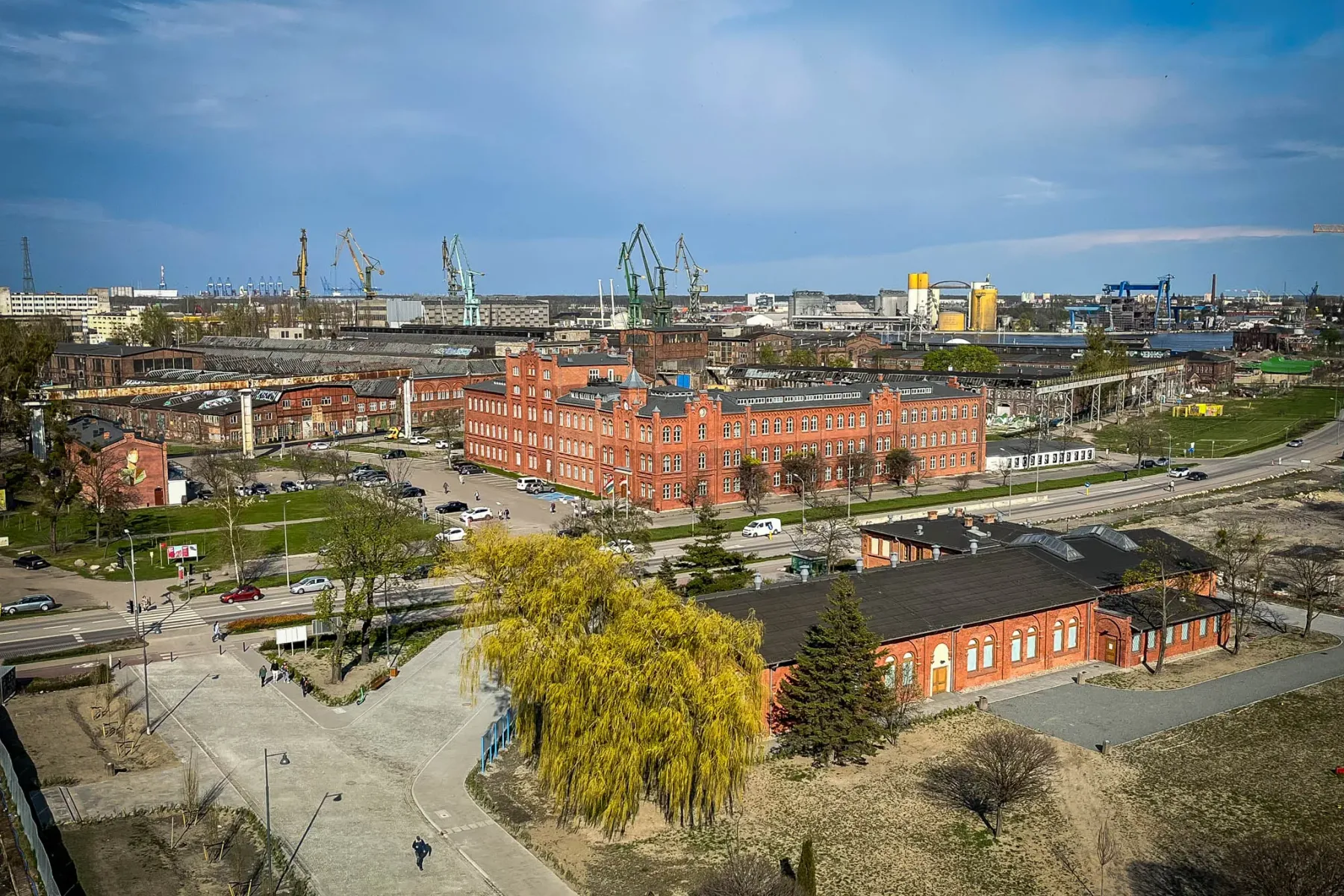 Luftaufnahme des Werftgebiets Danzig mit roten Backsteingebäuden, Straßen, Autos und Industriekränen unter blauem Himmel.