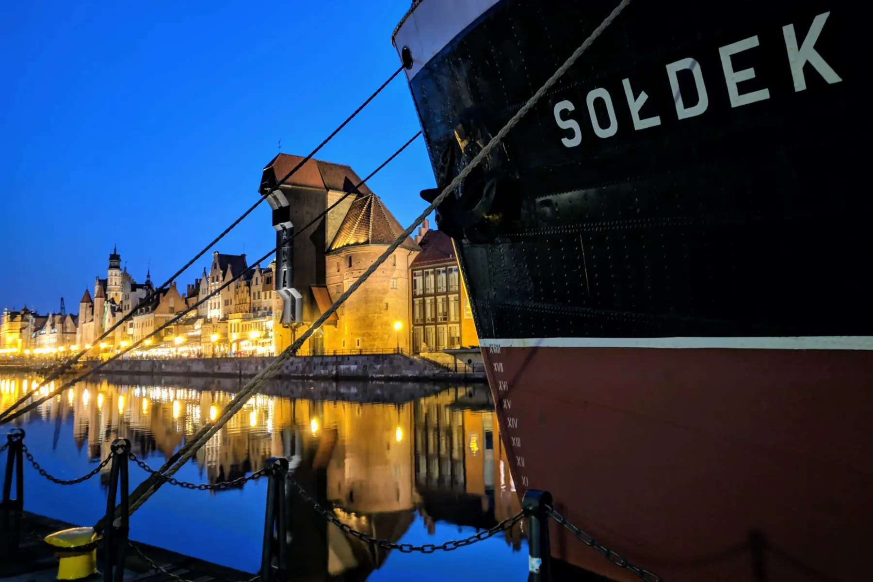Schwarzer Rumpf des Schiffs MS Sołdek am Kai, mit beleuchteten historischen Gebäuden und dem Kran Żuraw im Wasser gespiege...