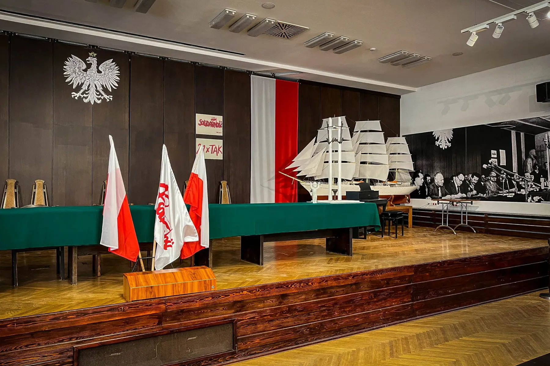 Konferenzbühne mit grünem Tisch, polnischen Flaggen, einem Segelschiffmodell und polnischem Adler an einer Holzwand.