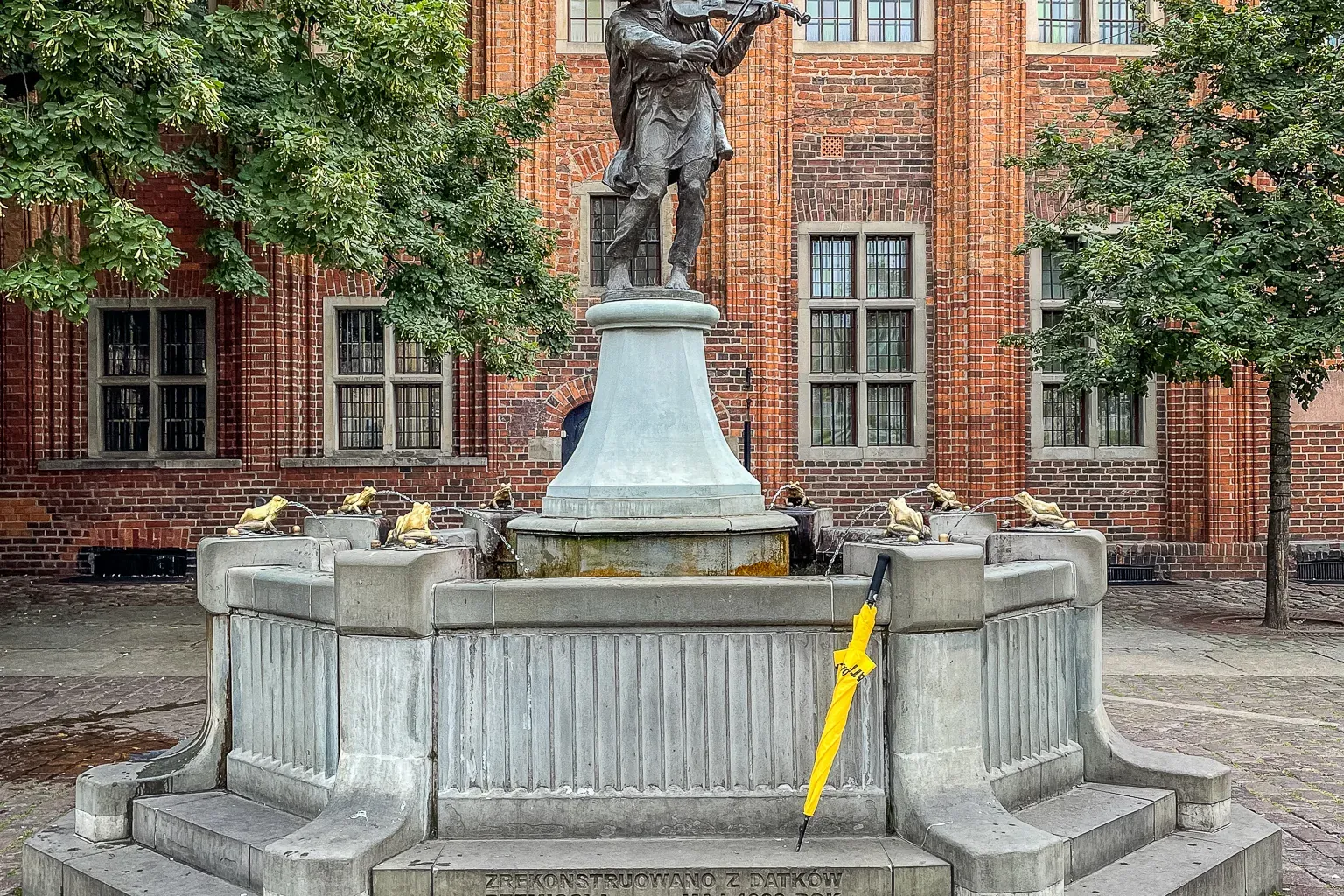 Bronzestatue eines Geigers, der Geige spielt, in einem Steinbrunnen mit Froschskulpturen vor einem roten Backsteingebäude....