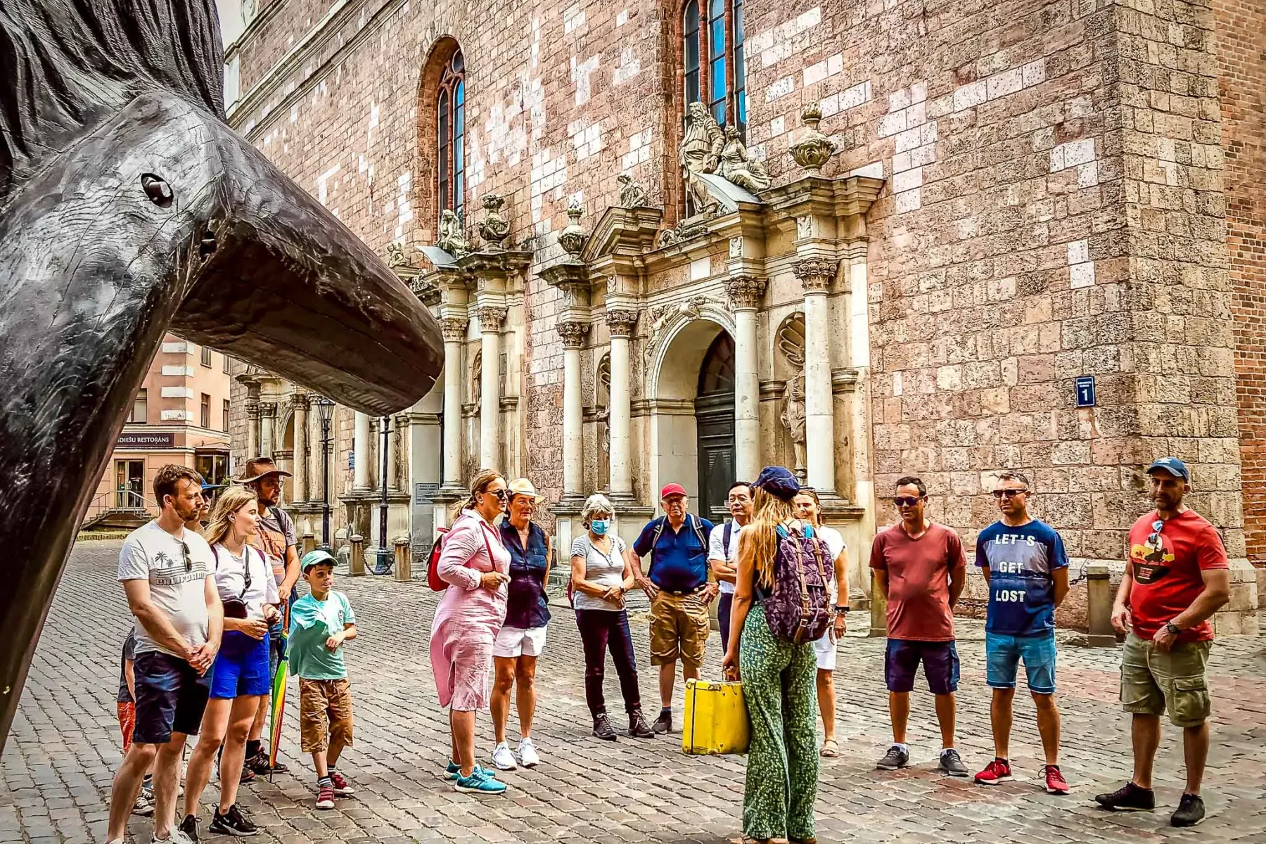 Reisegruppe hört einem Führer auf Kopfsteinpflasterstraße vor der Rigaer Petrikirche zu, mit einer Skulptur links.