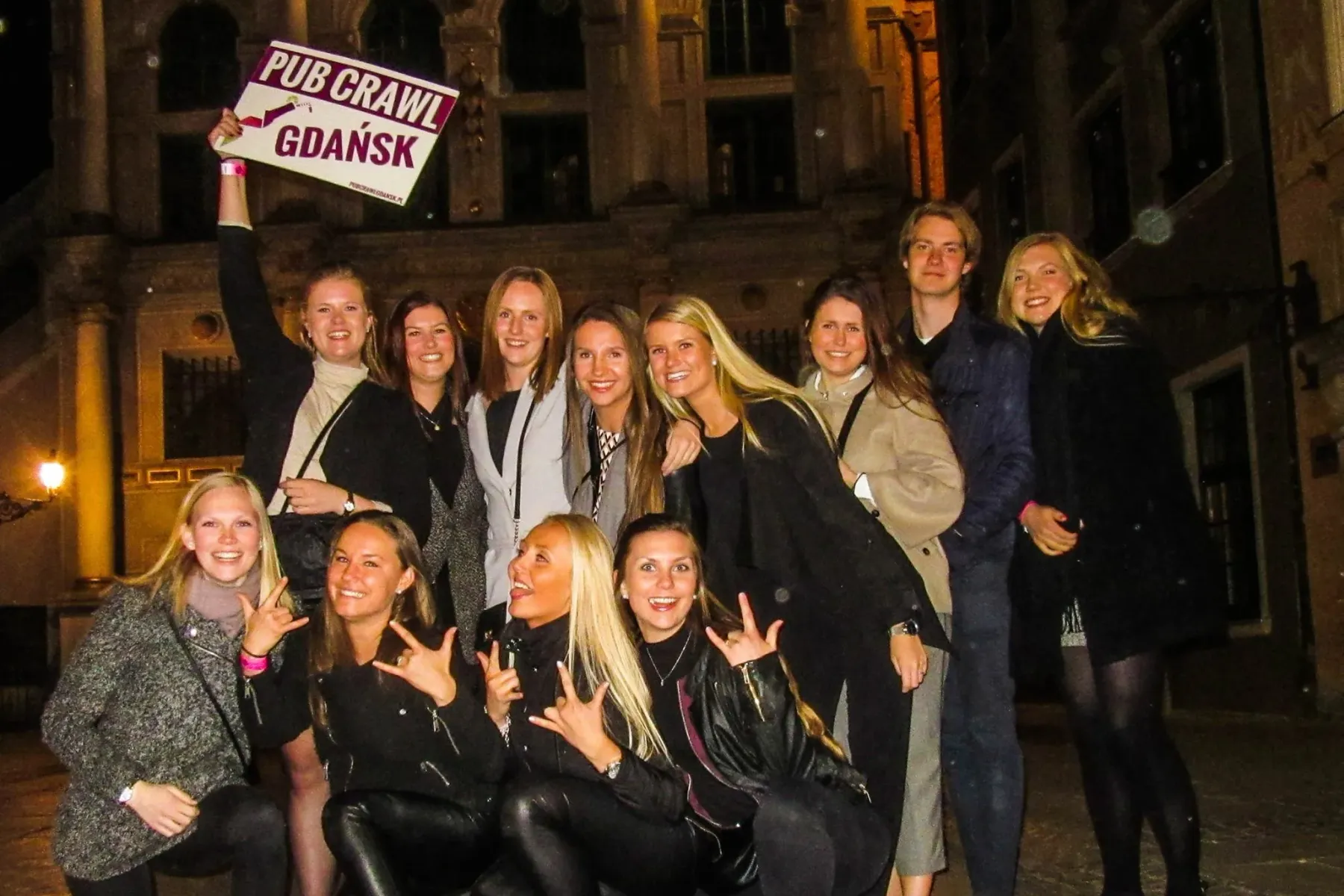 Eine Gruppe junger Erwachsener lächelt und posiert nachts vor einem historischen Gebäude. Eine Person hält ein Schild mit ...