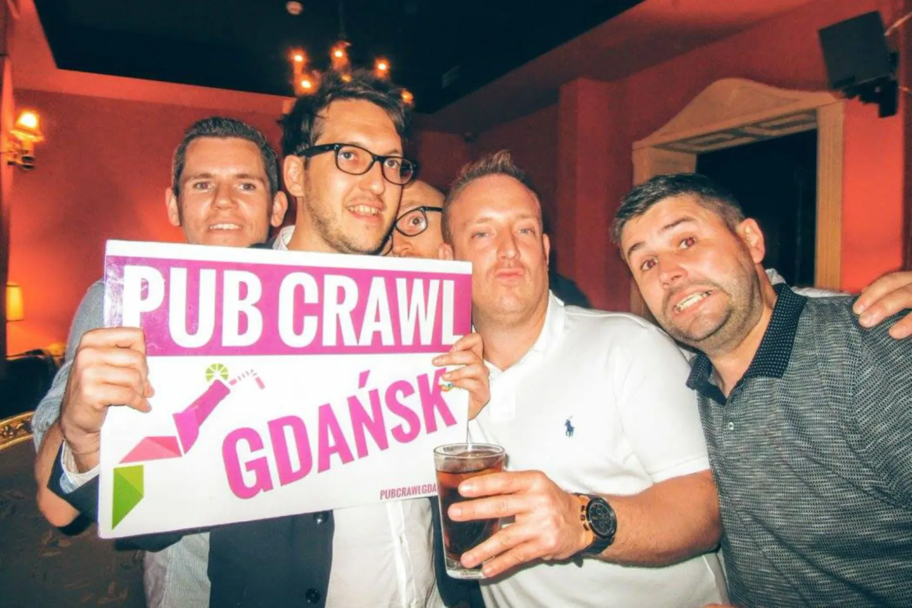 Vier Männer in einer schwach beleuchteten Bar, drei lächeln, einer hält ein weißes Schild mit der Aufschrift 'PUB CRAWL GD...