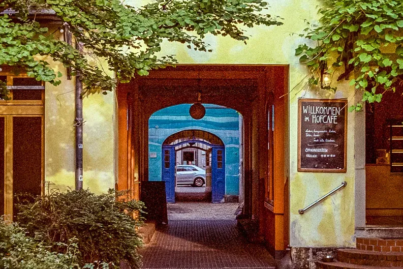 Gepflasterter Innenhof führt zu einem Torbogen, ein Gebäude mit gelben Wänden, Fenstern und einem 'Willkommen im Hofcafé' ...