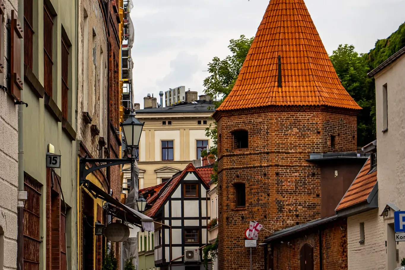 Kopfsteingasse mit historischen Gebäuden und einem runden Backsteinturm mit orangefarbenem Ziegeldach unter bewölktem Himmel.