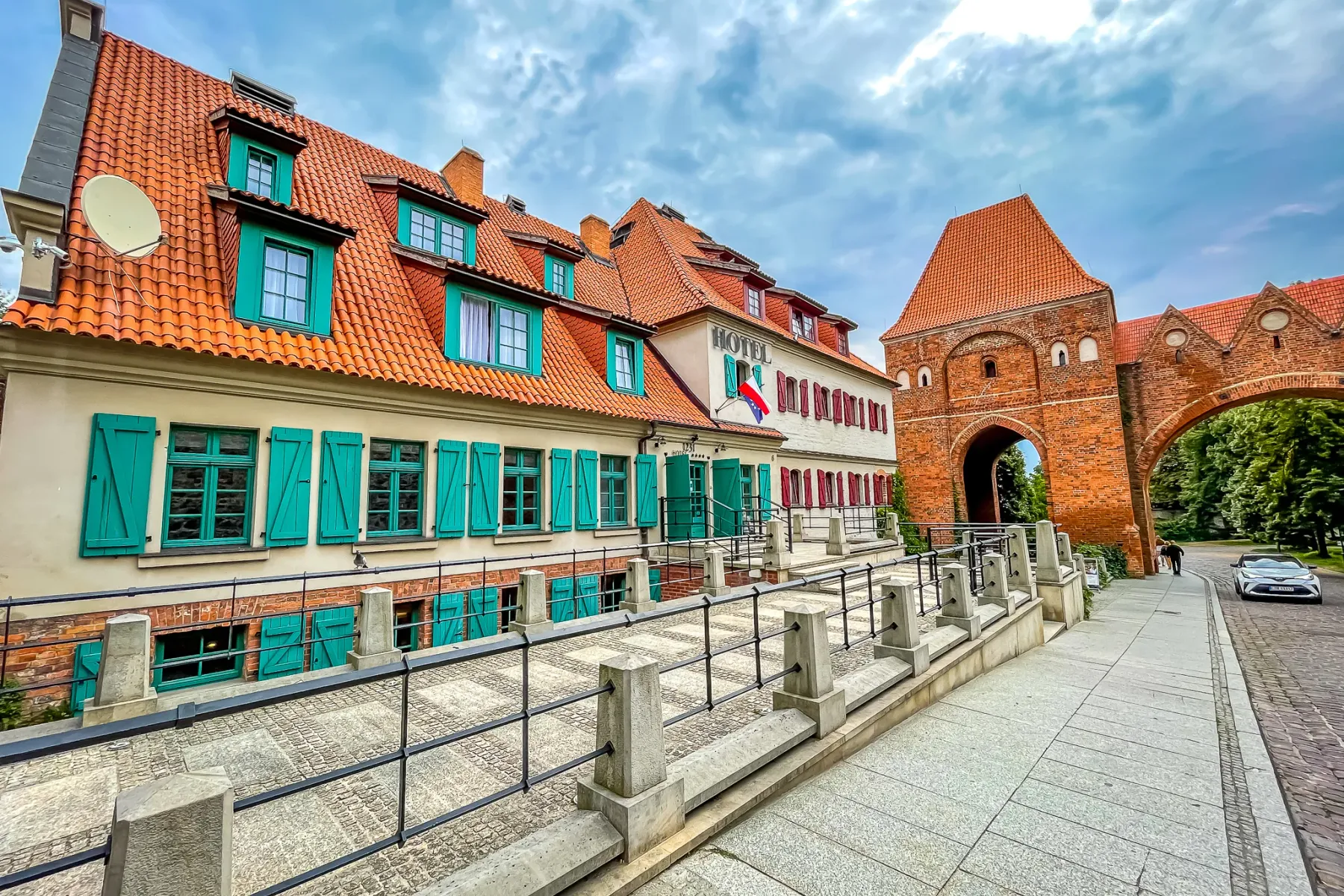 Historisches Hotel mit türkisfarbenen Fensterläden und rotem Ziegeldach neben einem Backsteintor und Steinweg unter bewölk...
