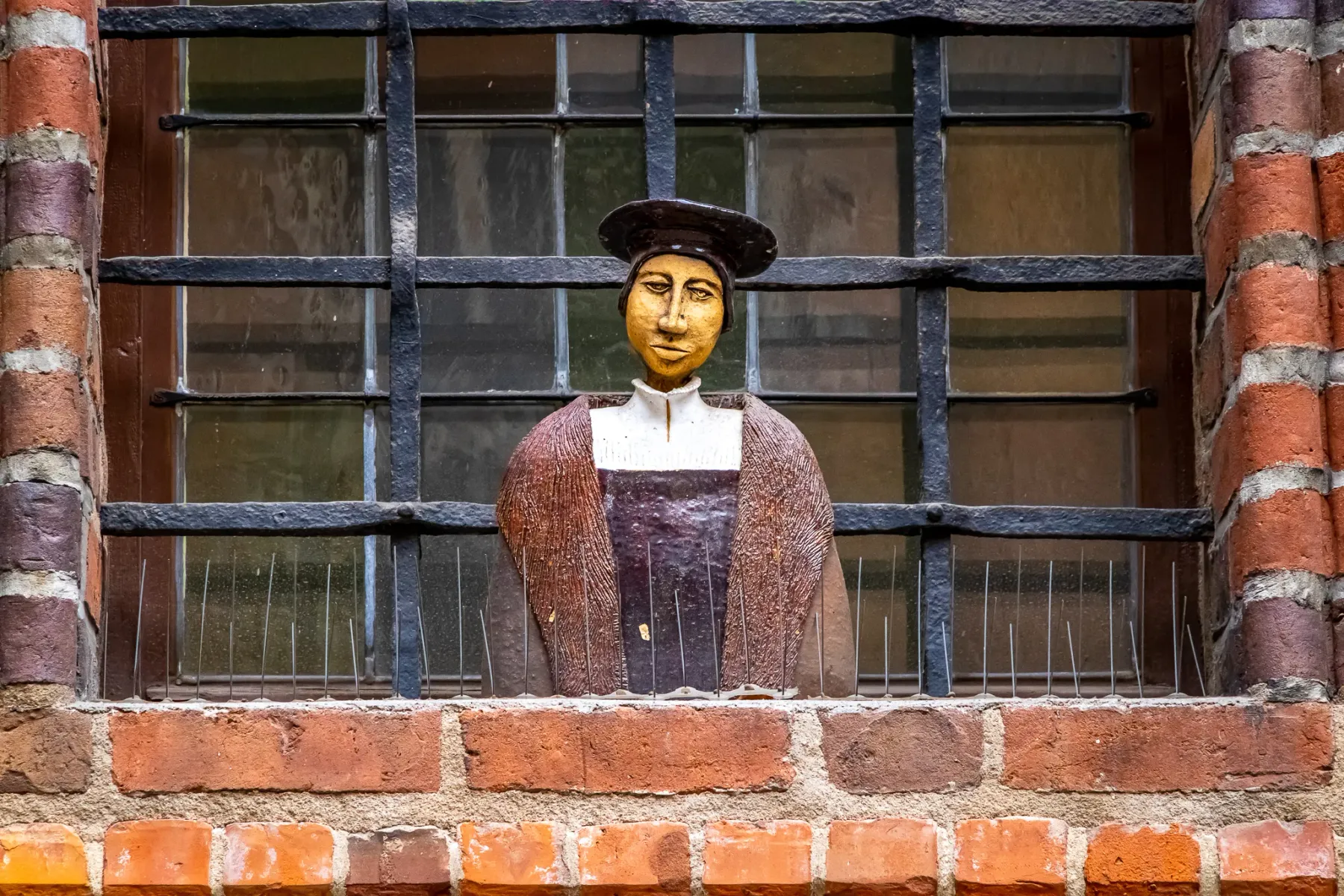 Stilisierte Figur mit Hut und historischer Kleidung in einem Ziegelsteinfenster mit Metallgitter. Unten sind Vogelabwehrsp...