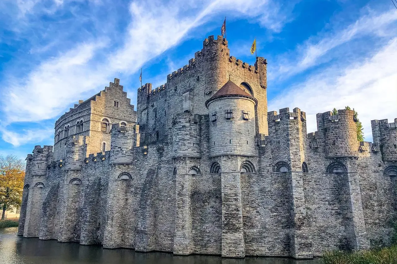 Castillo medieval de piedra con múltiples torres y almenas, rodeado por un foso oscuro bajo un cielo azul con nubes.