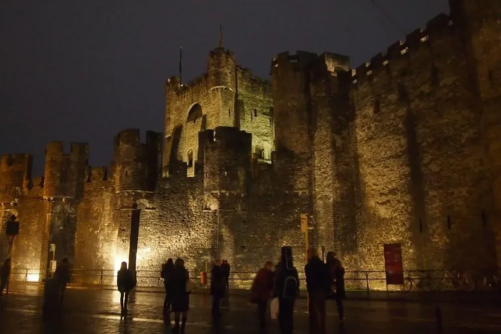 Gravensteen Castle in Ghent at night, illuminated by warm lights, with people standing on the wet street in front.