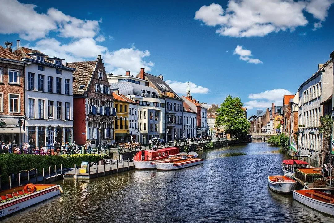 Edificios históricos con tejados a dos aguas bordean un soleado canal con barcos turísticos y gente en las orillas.