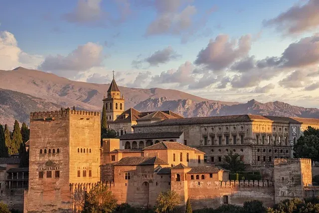 Historic Alhambra palace complex with a bell tower and surrounding walls, set against rocky mountains under a partly cloud...