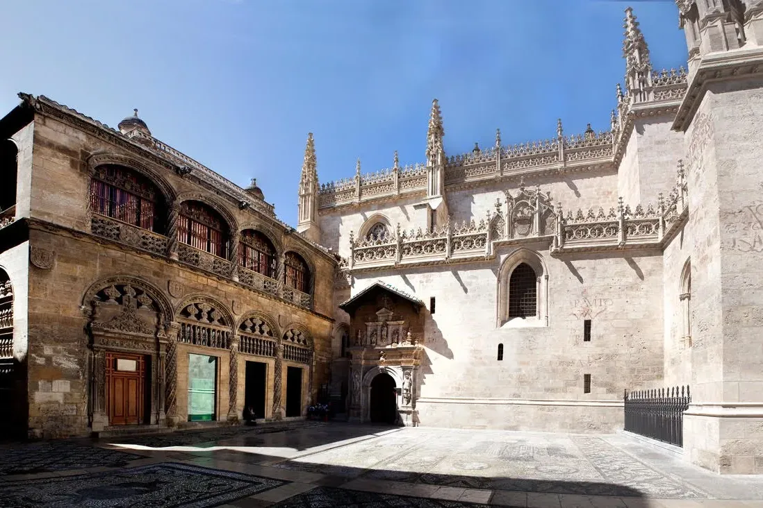 Ornate stone architecture of a chapel and an adjacent building in Granada, with a patterned courtyard under a blue sky.