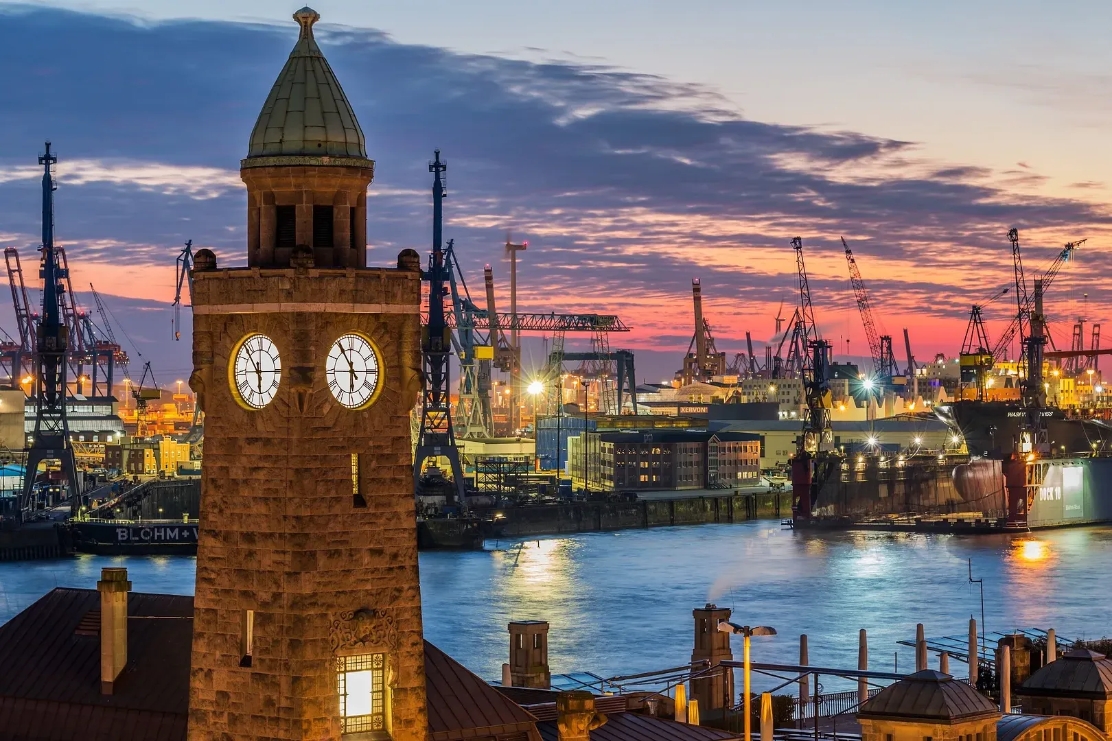 Torre del reloj de piedra con esferas iluminadas en Hamburgo. Al fondo, el puerto al anochecer con grúas, barcos y un ciel...