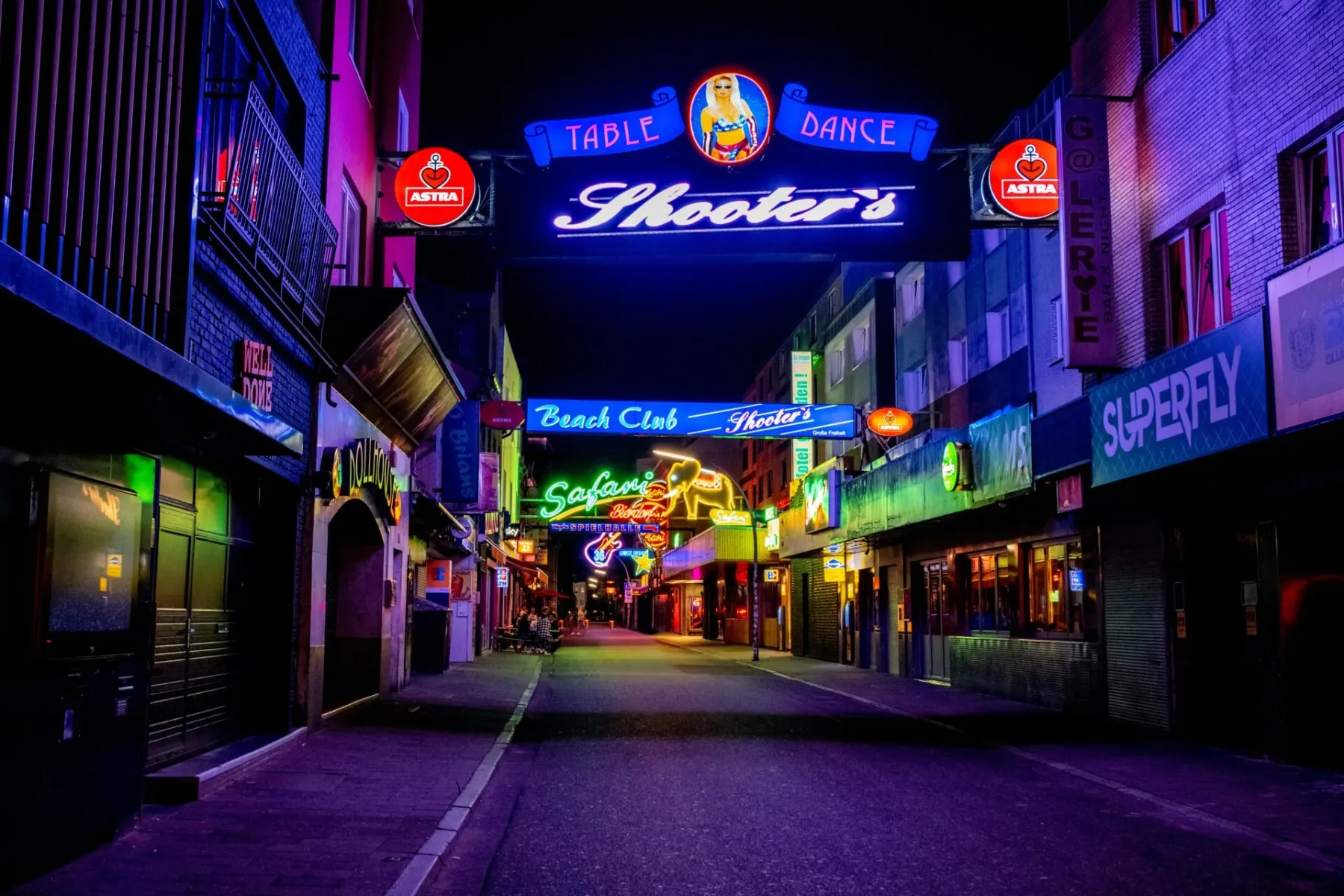 Calle de Hamburgo de noche, iluminada por letreros de neón de bares y clubes como "Shooter's" y "Safari" en tonos púrpura ...
