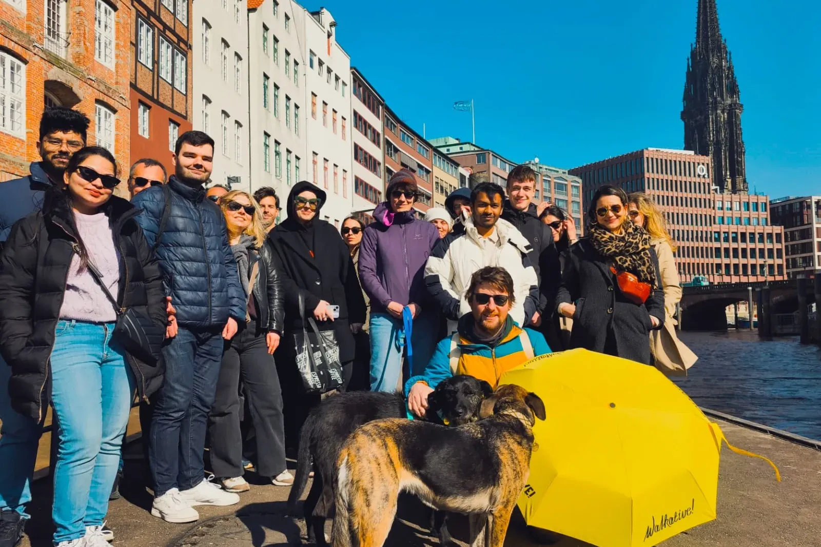 Grupo diverso de personas en un tour a pie junto a un canal en Hamburgo, con edificios históricos y la aguja de la iglesia...