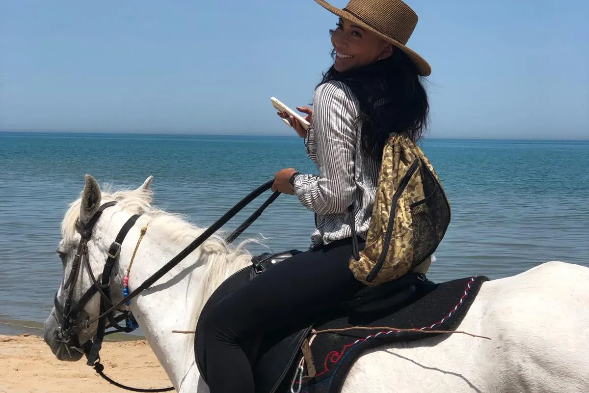 Mujer con sombrero de paja y mochila montando un caballo blanco en una playa de arena junto al mar azul tranquilo.