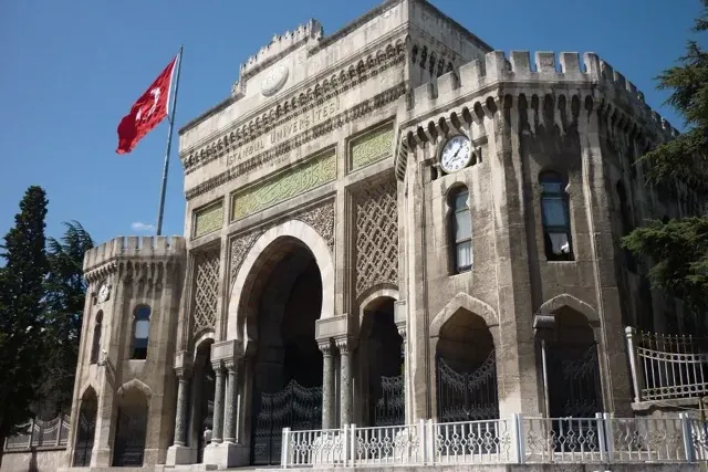 Majestuosa puerta principal de piedra de la Universidad de Estambul con una bandera turca ondeando y un reloj en una torre...