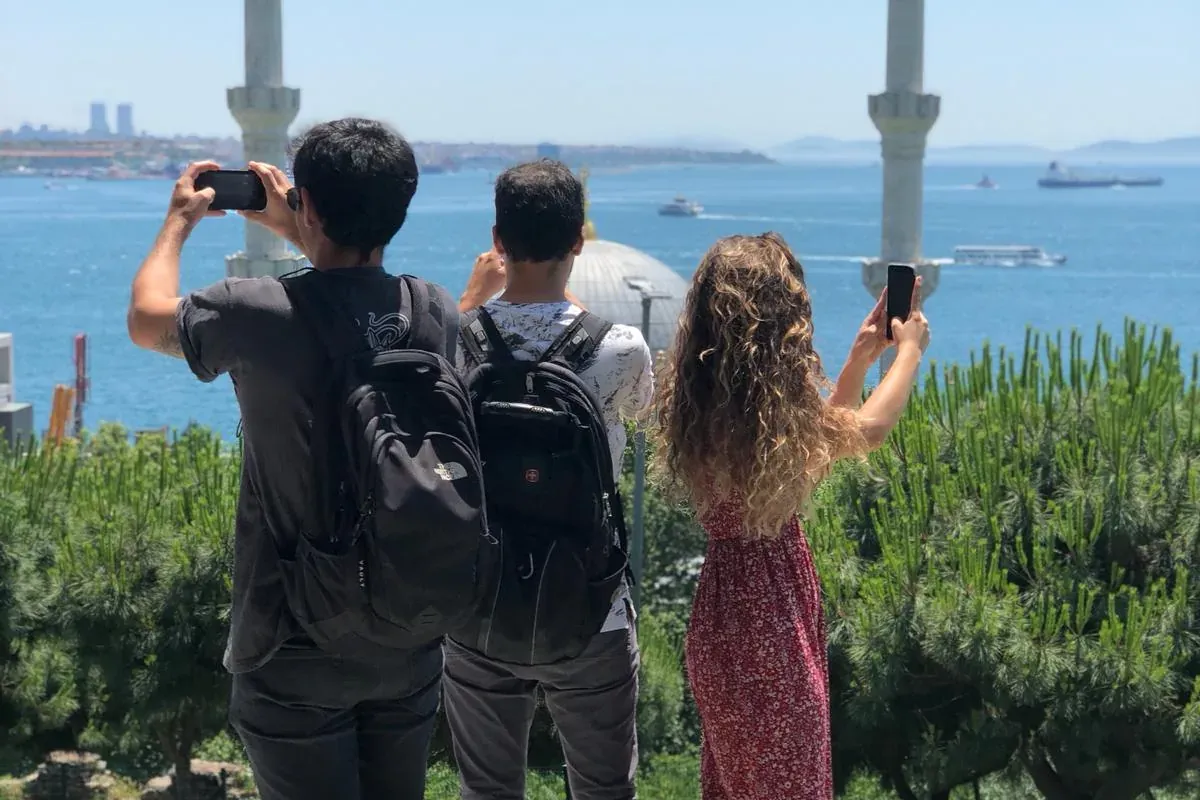 Tres personas fotografían el estrecho del Bósforo, con minaretes y una cúpula de mezquita al fondo, bajo un cielo azul claro.