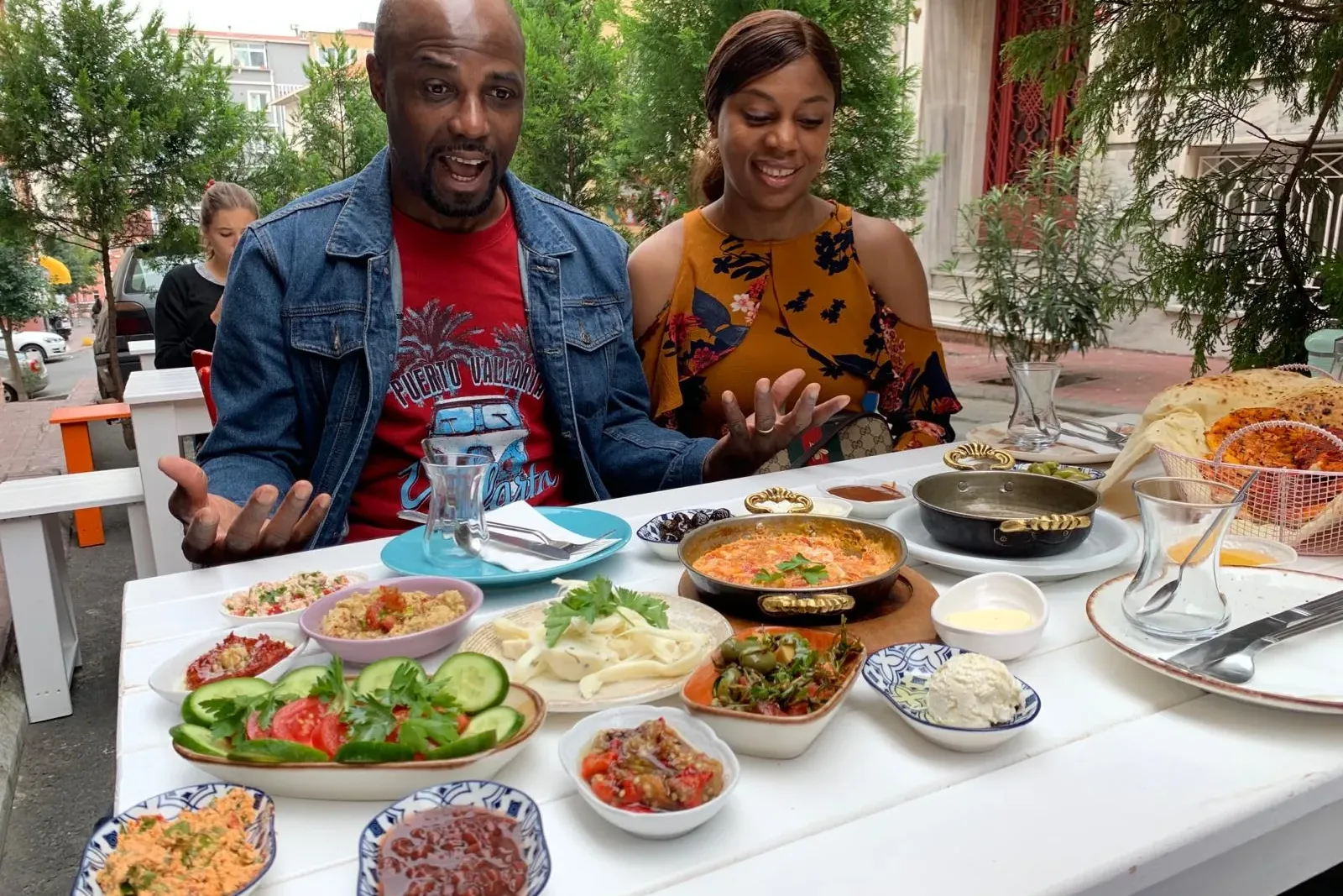Un hombre y una mujer en una mesa al aire libre repleta de una variedad de platos de desayuno turco, como huevos, meze y v...
