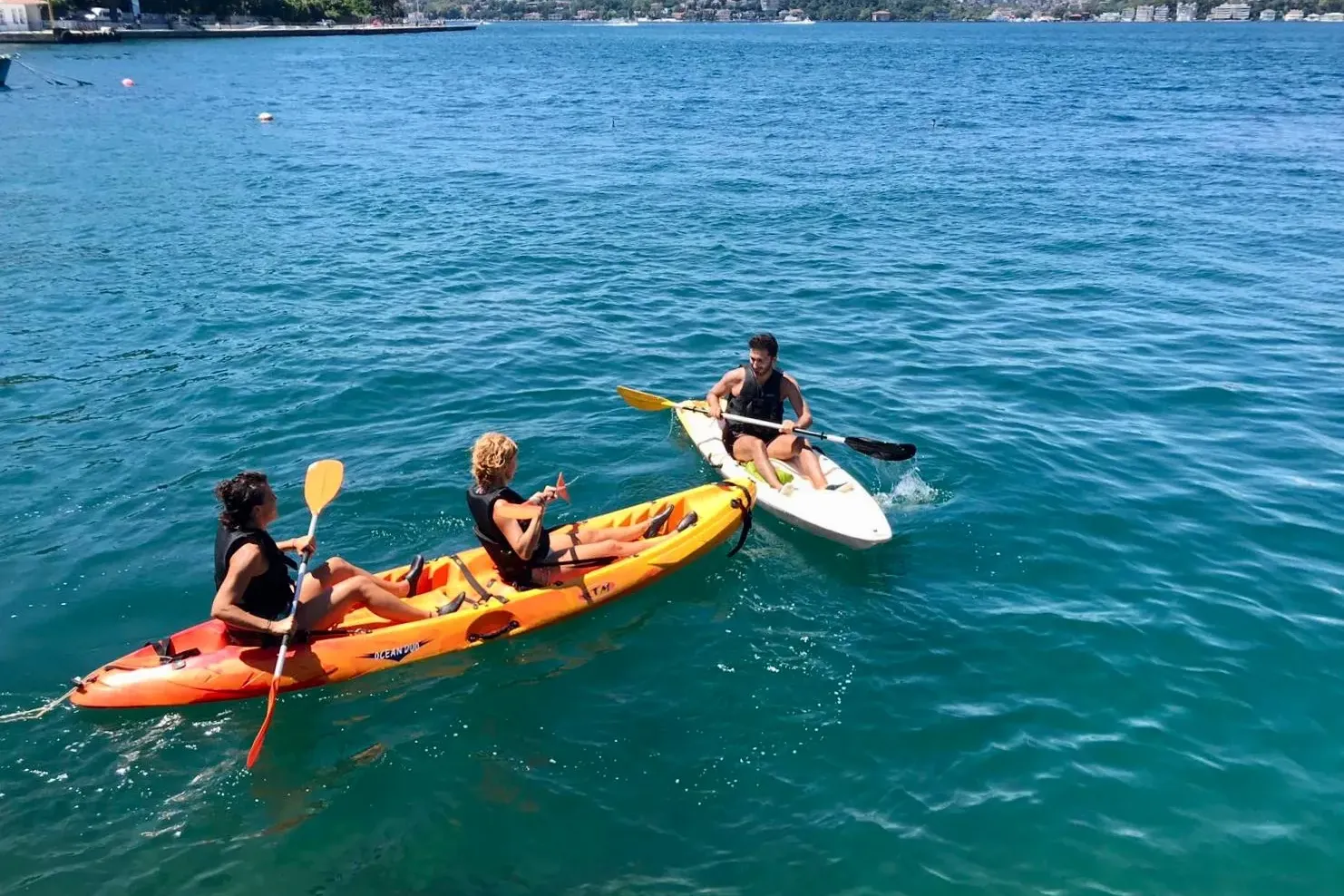 Tres personas con chalecos salvavidas en dos kayaks en agua azul. Dos en kayak naranja, una en kayak blanco. Orilla verde ...