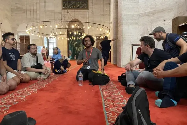 Un grupo de turistas sentado en una alfombra roja dentro de una gran mezquita con techos altos y arañas, escuchando a su g...