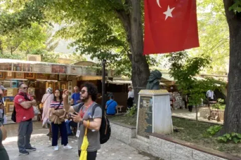 Guía turístico hablando a un grupo de turistas bajo árboles verdes. Una bandera turca cuelga de un árbol; hay una estatua ...