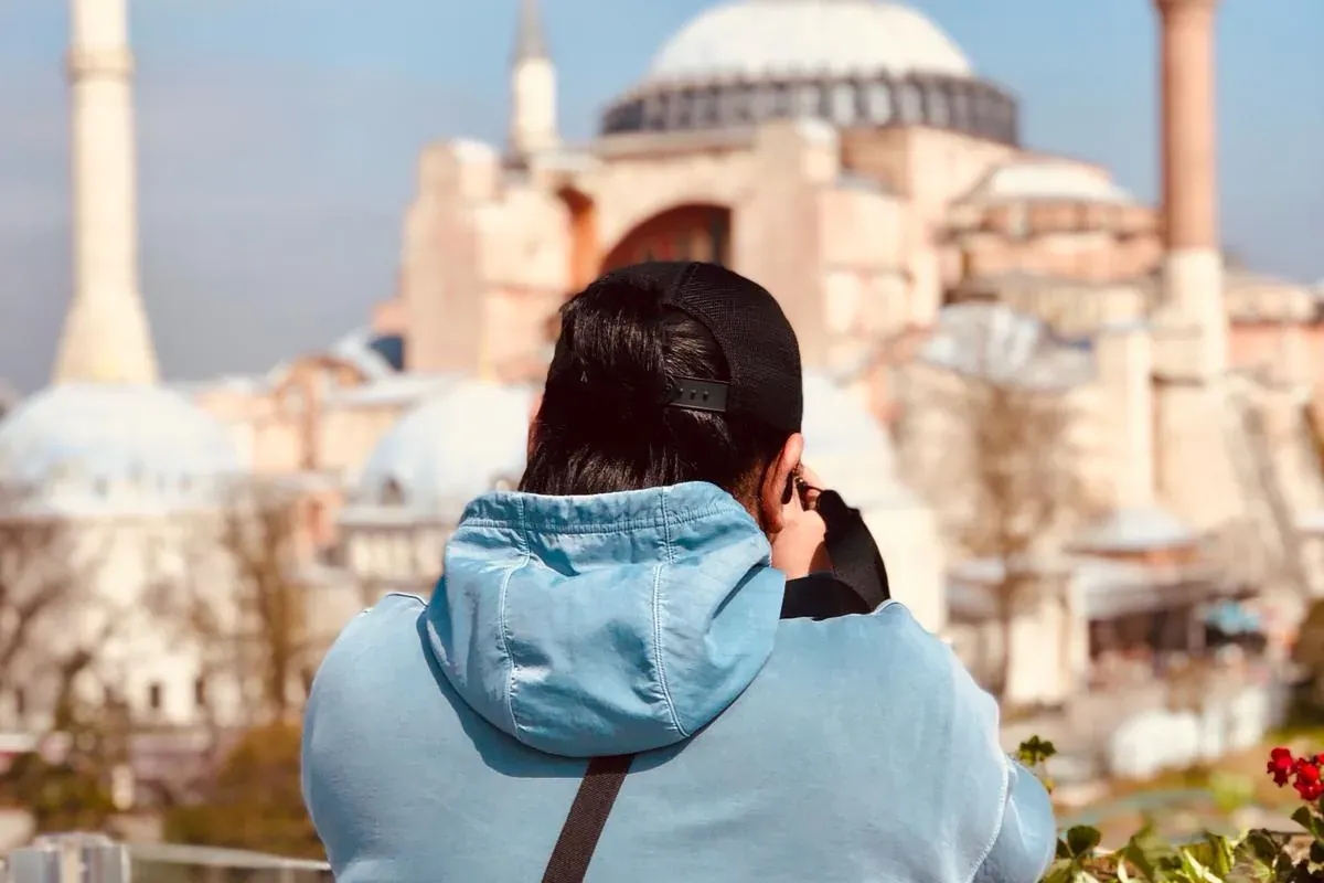 Hombre con sudadera azul y gorra negra observa Santa Sofía y sus minaretes bajo un cielo despejado desde una terraza.